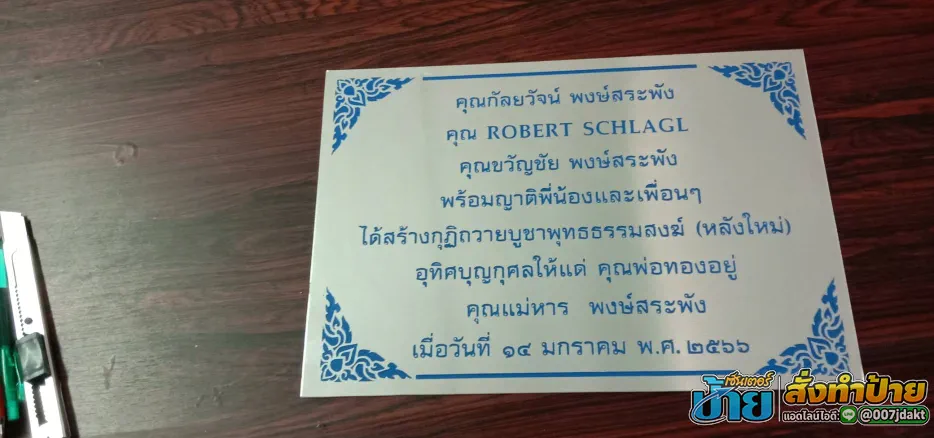 ป้ายรายชื่อผู้บริจาคสแตนเลสกัดกรดทรงสี่เหลี่ยมผืนผ้า พื้นสีเงิน ตัวอักษรสีน้ำเงิน จารึกชื่อผู้ร่วมสร้างกุฏิถวายบูชาพุทธธรรมสงฆ์ ตกแต่งลวดลายไทยสีน้ำเงินทั้งสี่มุม